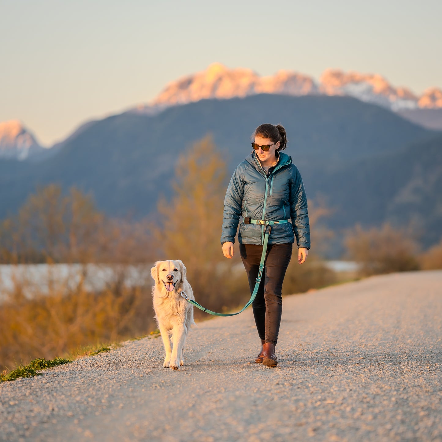 A woman walking a happy dog hands-free with a DogGo Collar, Leash and Belt in glacier teal at sunset.